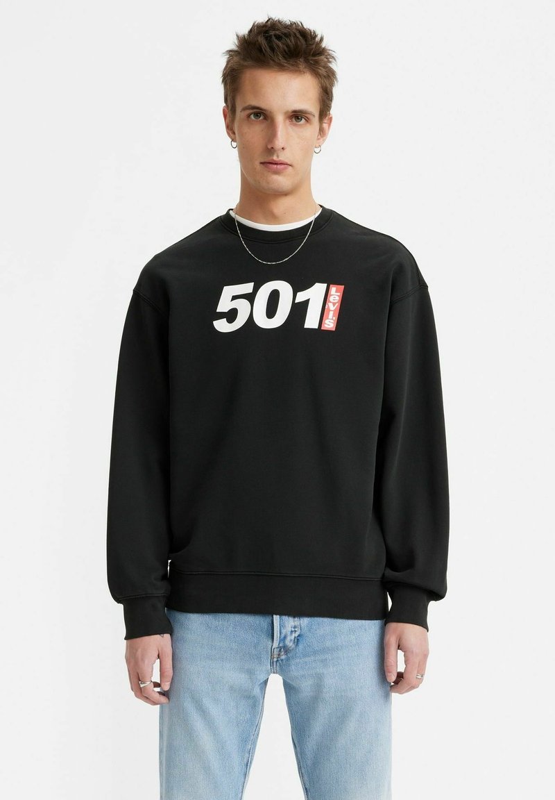 Zwarte sweatshirt met het "501" en "Levi's" logo in wit en rood, geribbelde manchetten en onderrand, ontspannen pasvorm, gedragen met lichtblauwe jeans.