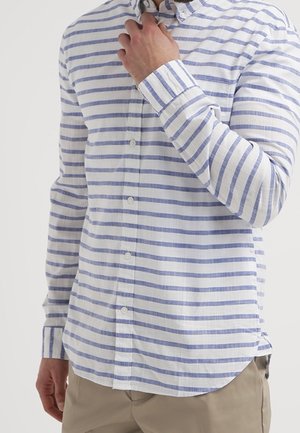 Mężczyzna noszący białą, długorękawową koszulę w jasnoniebieskie poziome paski, poprawiający kołnierz, w zestawieniu z beżowymi spodniami.