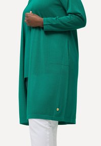 Zielona bluzka z długimi rękawami, wykonana z miękkiego materiału, z boczną kieszenią i złotym akcentem przy dołku. Noszona na białych spodniach.