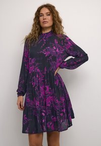 Kukkaistulosteen mekko tummansinisellä pohjalla, jossa on kirkkaanpurpleja kukkia, pitkät hihat, röyhelöhelma ja korkea kaulus kevyessä kankaassa.