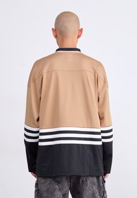 Άτομο με ξυρισμένο κεφάλι που φοράει μακρυμάνικο πουκάμισο σε μπεζ, μαύρο και άσπρο ριγέ σχέδιο και γκρι cargo παντελόνι, με θέα από πίσω σε απλό φόντο.