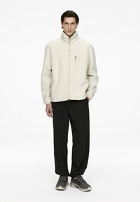 Fleece zip-up jacket in light beige with a high collar and chest pocket, paired with black pants and gray athletic shoes.