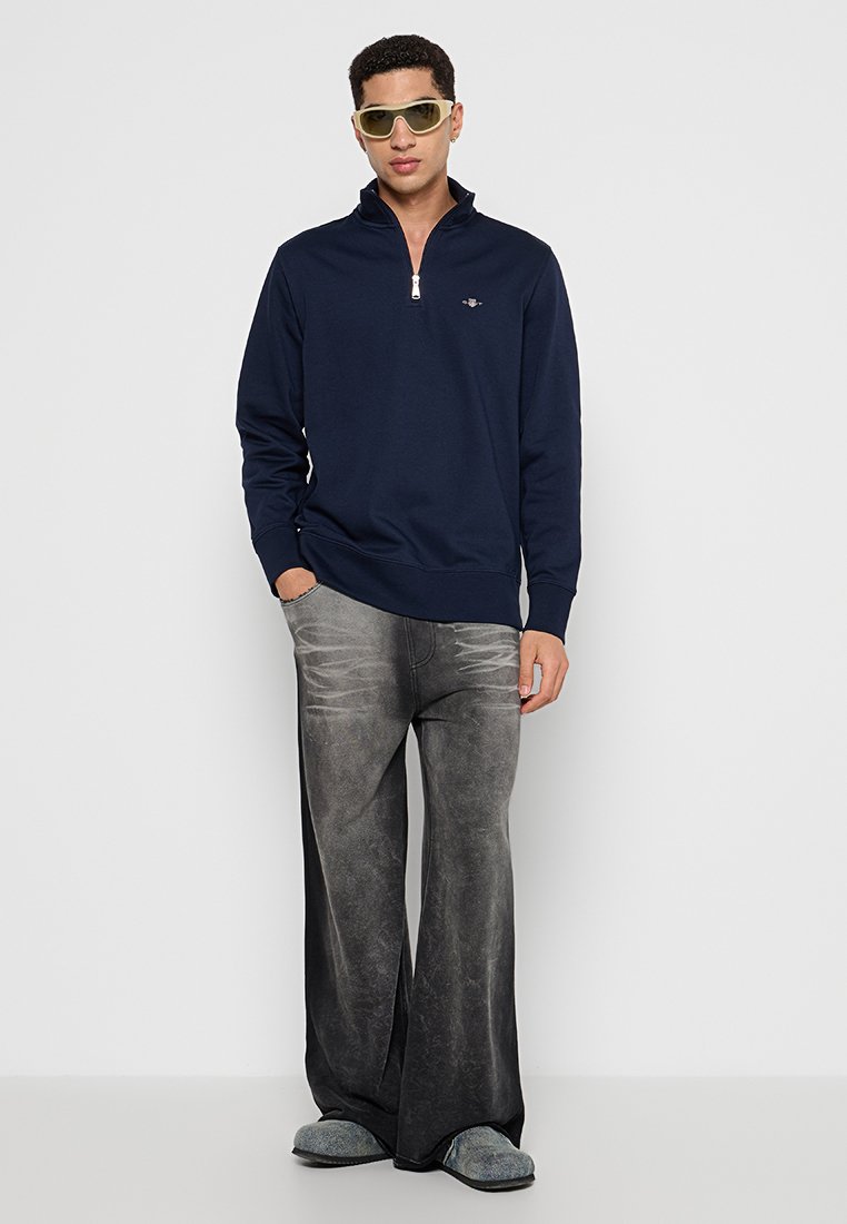 Junger Mann mit gelben Sonnenbrillen, marineblauem Zip-up-Pullover, weiten grauen abgenutzten Jeans und grauen Slippern, der vor einem weißen Hintergrund steht.