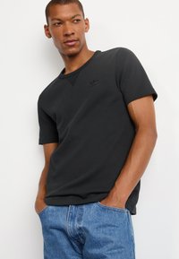 Must täheline must T-särk lühikeste varrukatega, tekstuurse viimistlusega, ümmarguse kaeluse ja väikese musta logoga rinnal. Kombineeritud siniste denim-pükstega.