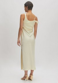 Gelbes Satinkleid mit lässigem Schnitt, verstellbaren Trägern und einem seitlichen Schlitz. Das Modell trägt beige High Heels. Glatte Textur.