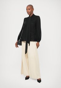 Juoda modeliuota palaidinė su ilgomis, pūkuotomis rankovėmis ir priekine užrišimo detalė. Šviesiai rudos plačiakelnės kelnės ir juodi smailiakulniai bateliai. Maža petinė rankinė.