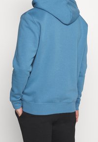 Blå bomullshuvtröja med känguruficka, ribbade ärmslut och fåll. Har en dragsko i huvan och en slät, mjuk textur.