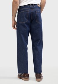 Men's blue denim jeans with a straight fit, visible seams, two back pockets, frayed hems, and brown leather shoes.