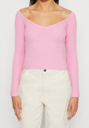 Pull en maille rose à épaule dénudée, à manches longues et avec des détails texturés sur le buste, associé à un pantalon de couleur claire.