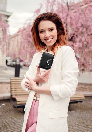 Black leather wallet with a silver hardware accent. Features a rectangular shape and a logo, held by a person in a white blazer and pink pants.