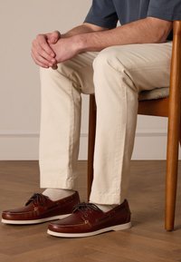 Scarpe da barca in pelle marrone con suole bianche, parte superiore testurizzata e lacci. Indossate con pantaloni color crema, seduto su una sedia di legno.