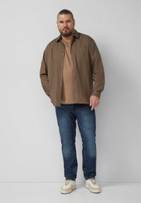 Brown printed button-up shirt over a beige t-shirt, dark blue jeans, and white sneakers. The shirt features a subtle pattern and a relaxed fit.