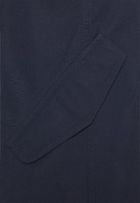 Tela azul marino con una textura suave, con un solapa de bolsillo diagonal y costuras en contraste. El diseño enfatiza líneas limpias y funcionalidad.