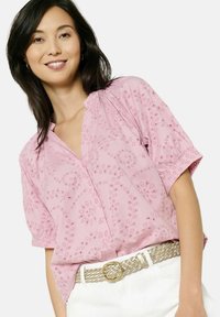 Rozā īsām piedurknēm krekls ar ziedu izšuvumu, V veida kakla izgriezumu un pogu detaļām, kombinēts ar austu jostu un baltām biksēm.