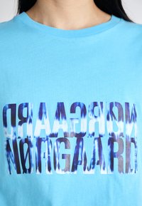 Light blue cotton t-shirt featuring large, distorted text in dark blue and white. Crew neck design with short sleeves.