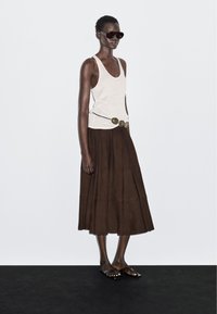 Vrouw in een witte tanktop, bruine midi-rok met een grote ronde gesp, donkere zonnebril en zwarte sandalen, staand op een zwarte vloer.