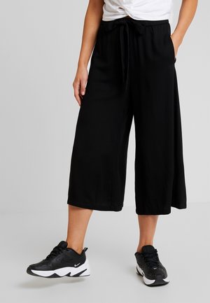 Persoon die zwarte wijde cropped broek met een strikband in de taille en zwart-witte sportschoenen draagt, staand op een grijze vloer.