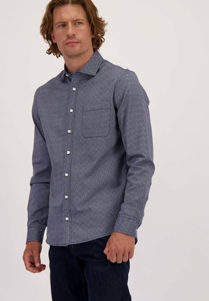 Man in een blauw gestreept overhemd met lange mouwen en een borstzak, gecombineerd met donkere jeans, staande tegen een effen achtergrond.