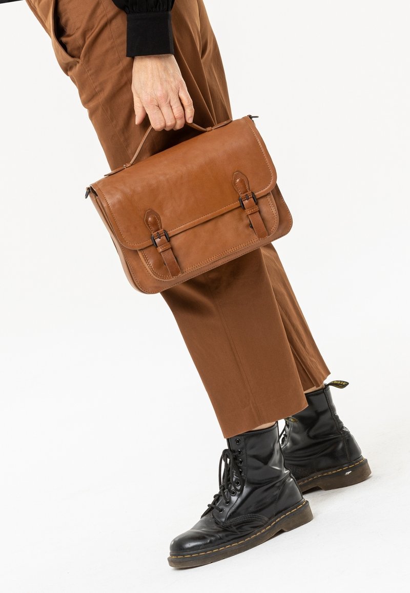 Personne tenant une sacoche en cuir marron par la poignée, portant un pantalon marron et des bottes noires à lacets avec surpiqûres jaunes.