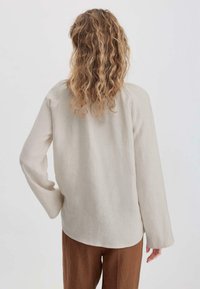 Camicia in lino beige con maniche lunghe, orlo arrotondato e vestibilità rilassata. Abbinata a pantaloni marroni e larghi. Texture naturale visibile.