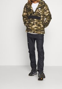 Sudadera de forro polar con estampado de camuflaje verde, media cremallera, bolsillo frontal negro y mangas largas, combinada con pantalones y calzado negro.