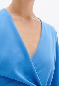 Blå inslagstopp med djup V-urringning, mjuk bomullstyg och slät yta med rynkade detaljer vid midjan.