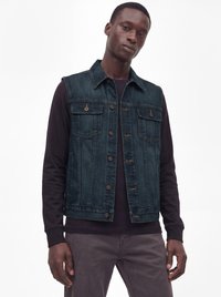 Junger Mann, der eine dunkelblaue Jeansweste über einem schwarzen Langarmshirt und grauen Hosen trägt, steht vor einem schlichten weißen Hintergrund.