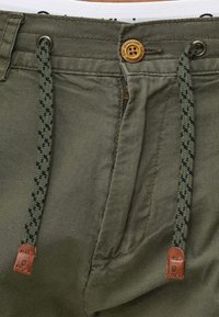 Khaki broek met een ritssluiting, verstelbare geweven trekkoord met een donkergroen en zwart patroon, en een bruine leren accent nabij de zoom.