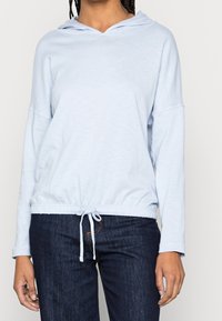 Ljust blå huvtröja med dragsko i midjan, mjukt tyg, sänkta axlar och långa ärmar, tillsammans med mörka denimjeans.