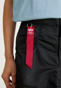 Crveni tkaninski privjesak za ključeve s logotipom "Alpha Industries Inc U.S.A." pričvršćen za džep crnih hlača, pored ruke osobe i svijetloplave košulje.
