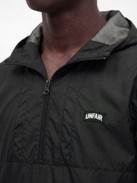 Primo piano di una persona che indossa una giacca nera con zip e una fodera testurizzata nel cappuccio, con una toppa etichettata "UNFAIR" sul petto.