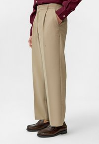 Beige, geplooide broeken met verticale streepjes; gemaakt van een gestructureerde stof. Draagt een paar donkerbruine instapschoenen met een gladde afwerking.
