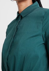 Camicia verde teal con colletto classico, tessuto liscio e vestibilità strutturata. Presenta un leggero lucido e dettagli di cucitura puliti.