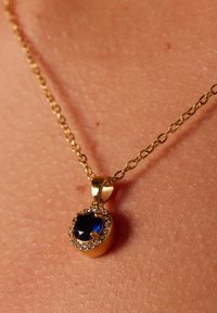 Gouden ketting met een blauwe edelsteen hanger, omringd door kleine heldere stenen, hangend aan een delicate ketting tegen de huidtextuur.