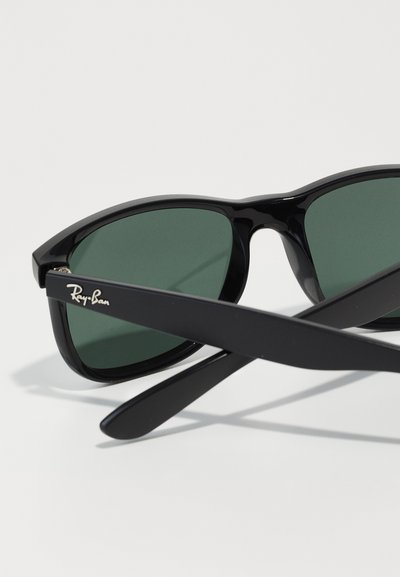 Lunettes de soleil rectangulaires noires avec des verres verts, présentant une monture et des branches brillantes. Logo Ray-Ban gravé sur le côté.