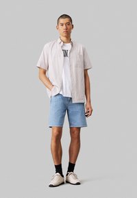Camicia a maniche corte a righe in beige chiaro e bianco, indossata sopra una T-shirt bianca con testo, abbinata a pantaloni corti in denim blu e sneakers beige.