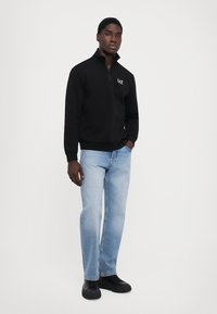 Sweatshirt negru cu guler tip fermoar și logo-ul EA7, asortat cu blugi drepti de un albastru deschis și adidași negri. Design simplu și stil casual.