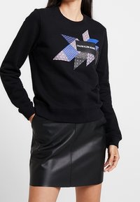 Svart sweatshirt med en geometrisk lapptäckesdesign i olika nyanser av blått och lila, med "Calvin Klein Jeans"-logotypen framtill.