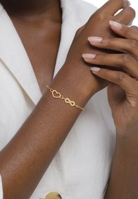 Guldband med ett hjärta och oändlighetssymbol, med en delikat kedjedesign. Armbandet bärs på en brun arm.