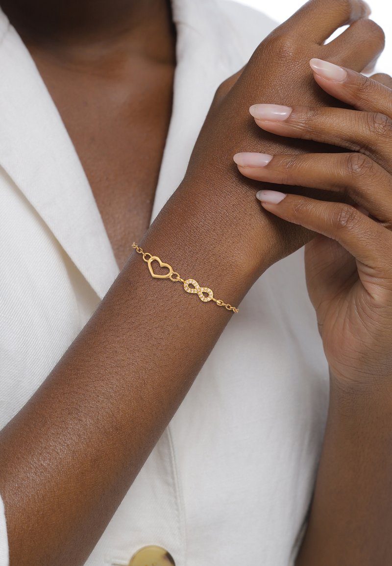 Guldband med ett hjärta och oändlighetssymbol, med en delikat kedjedesign. Armbandet bärs på en brun arm.