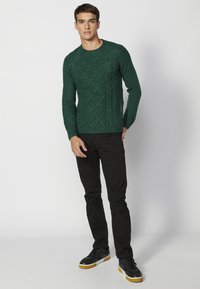 Groene gebreide trui met een gestructureerd diamantpatroon, ronde halslijn en ribgebreide manchetten. Gecombineerd met zwarte broek en zwarte sneakers.