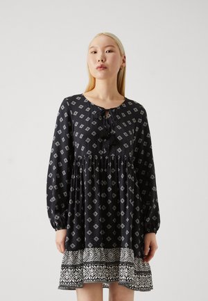 Robe noire avec des motifs en diamants, manches longues et taille froncée. Présente un col cintré avec un nœud et un ourlet à motifs blancs.