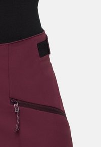 Gros plan d'une jupe bordeaux avec poche zippée diagonale et fermeture velcro noire à la taille, portée avec un haut noir.
