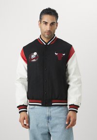 Melnbalts sporta žakets ar sarkaniem akcentiem, kuram ir bullim līdzīga emblēma un izšuvums "5x Čempioni", ar pogām aizdari un svītrainām piedurknēm.