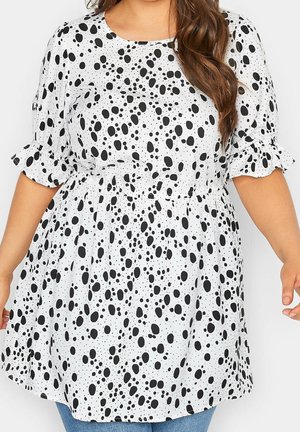 Femme portant une robe tunique blanche à pois noirs irréguliers, manches bouffantes et taille élastique, vue du cou jusqu'aux cuisses.