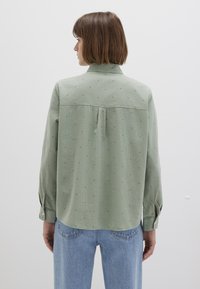 Grüne Button-Up-Hemd mit langen Ärmeln, das dezente, mehrfarbige Punktmuster aufweist, eine Rückenpasse hat und einen lockeren Schnitt bietet. Kombiniert mit hellblauen Jeans.
