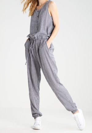 Gestreepte jumpsuit in grijs en wit, met een mouwloos ontwerp, knoopsluiting aan de voorkant, elastische taille en taps toelopende pijpen; gecombineerd met witte sneakers.