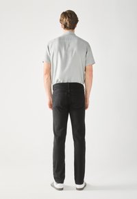 Hombre de pie frente a una pared blanca, vistiendo una camiseta de manga corta gris claro, jeans negros y zapatillas blancas con rayas negras.