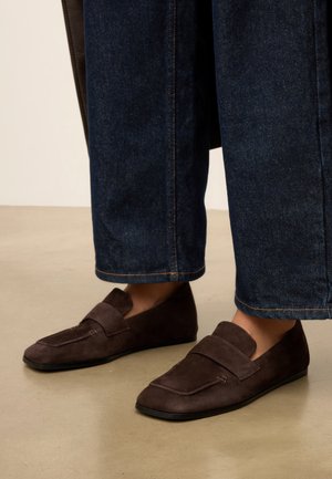 Bruine suède loafers gedragen met donkerblauwe cropped jeans op een lichtbeige vloerachtergrond.