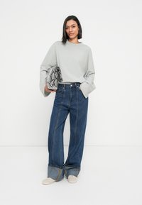 Φαρδιά γκρι ανοιχτόχρωμη μπλούζα με μακριά μανίκια, συνδυασμένη με μπλε τζιν παντελόνι φαρδιάς γραμμής με ραφές μπροστά και διακριτική φουσκωτή λεπτομέρεια στο τελείωμα. Κρατάει μια υφασμάτινη τσάντα με ασπρόμαυρο σχέδιο φιδιού.